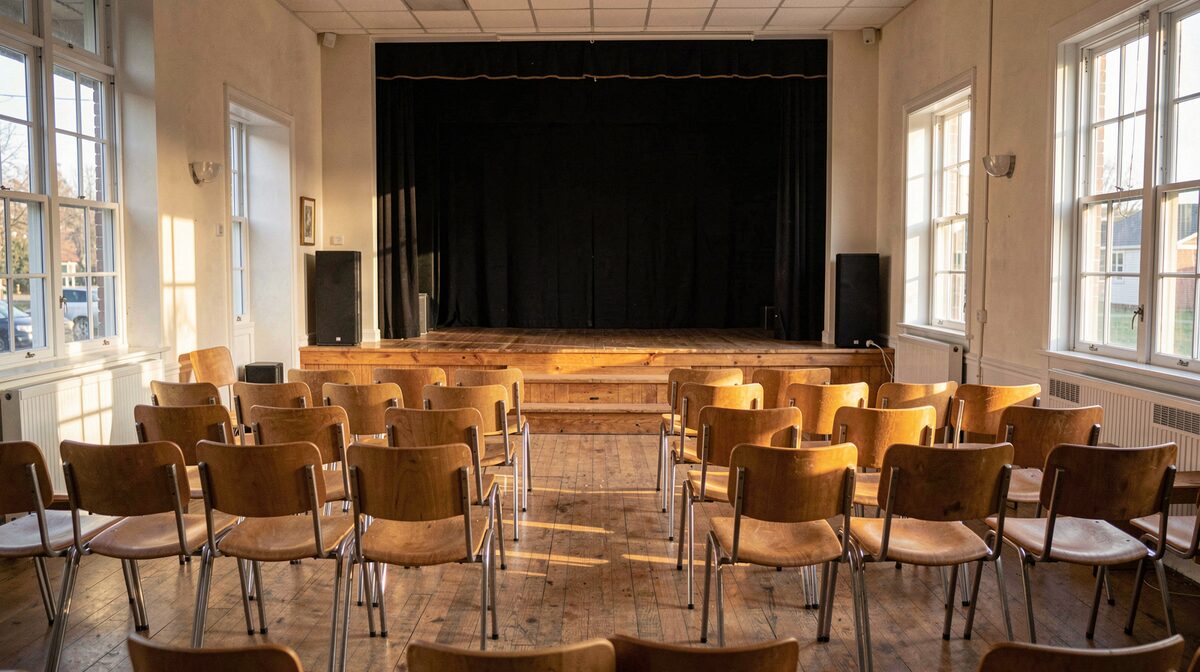 Une salle de répétition associative en fin d'après-midi, chaises alignées face à une petite scène en bois, lumière naturelle par les fenêtres, cadrage large depuis le fond de la salle.